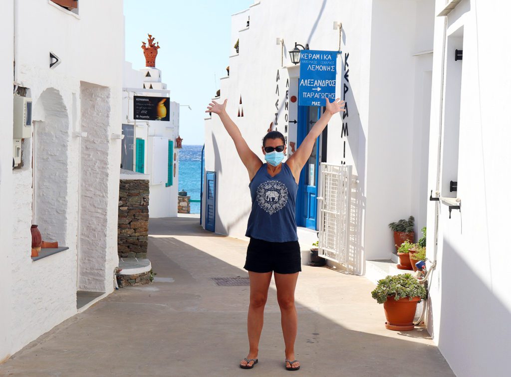 Robyn holding up both arms, with a face mask on, in a tiny alley on the island of Sifnos. You can see the turquoise waters behind the white washed buildings.