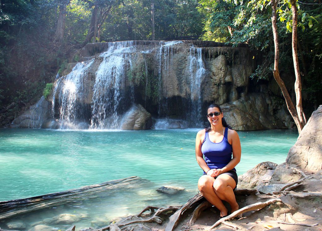 Robyn sitting on large boulders in front of the Erawan Waterfall in Thailand. She is always smiling, despite the pain.
