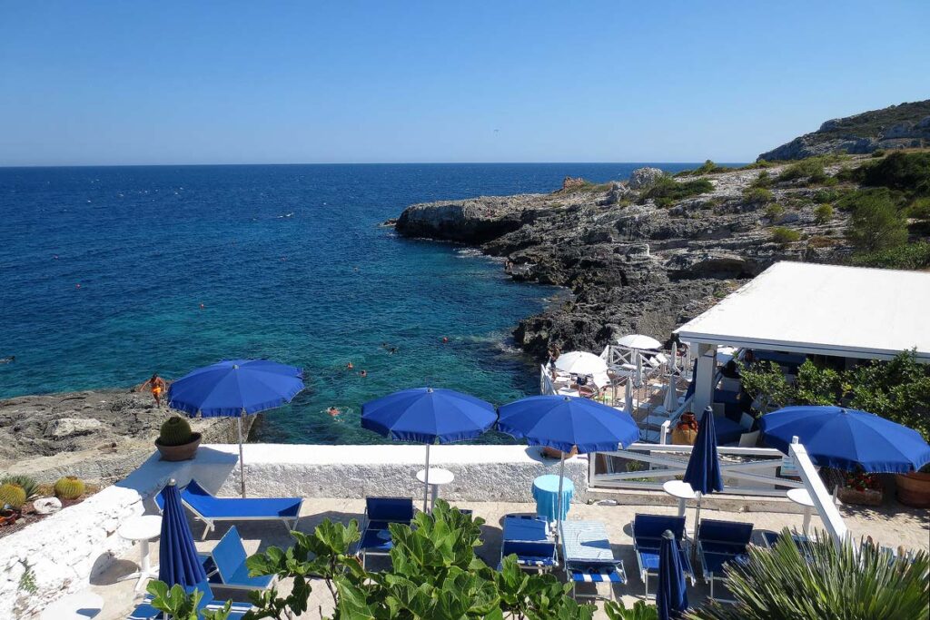 A waterfront spot in Marina di Novaglie in Puglia with blue sun loungers and umbrellas set up close to the cobalt water.