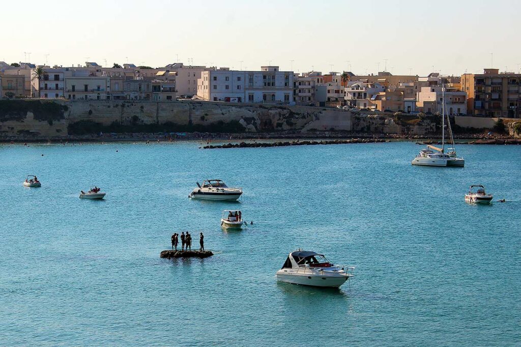 A beautiful view of the bay near Otranto. The walled city is in the distance and several boats are in the water as the sun is beginning to set.