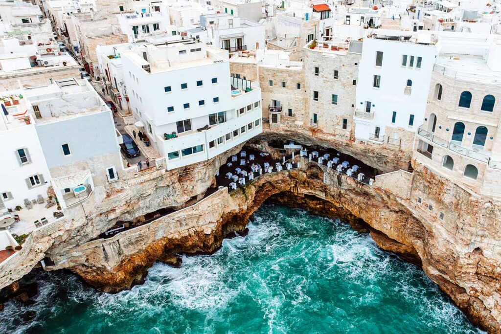 A bird's eye view of the stunning cliffs and white buildings of Polignano a Mare, one of the prettiest towns in Puglia to visit. there is a restaurants with white tables and chairs cut into the rockface above the sea.