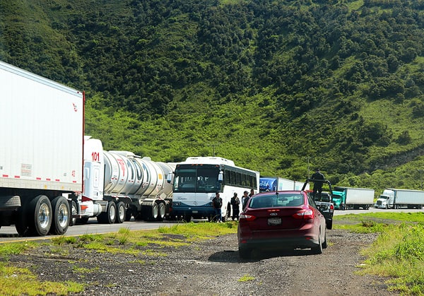 A line up of cars, busses and semis on the side of the road. A police truck is escorting a vehicle on a side dirt road. If you are caught in a long line when driving in Mexico, be prepared to wait a long time before resuming your trip.