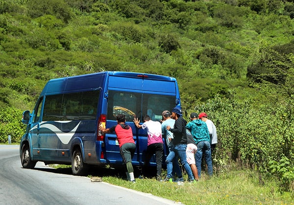 A group of men pushing a small blue van on the side of a green, hilly mountain road.