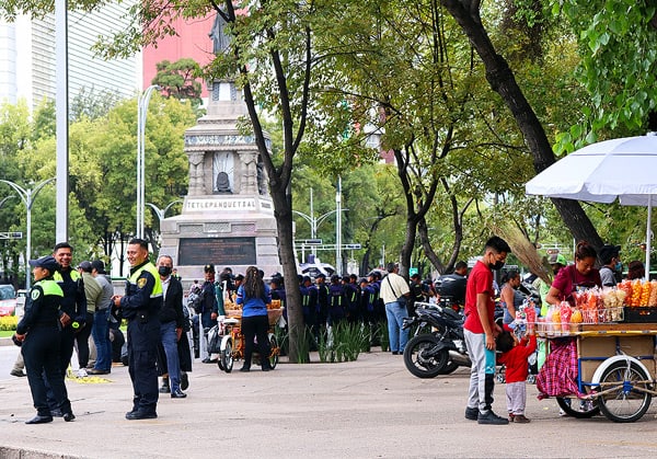 A group of 4 police standing near a vendor selling produce from his cart in a park in Mexico City. There are green trees and people walking around.