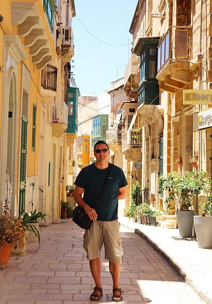 Gavin smiling in a narrow street. Tall sandstone buildings and colorful balconies surround him.