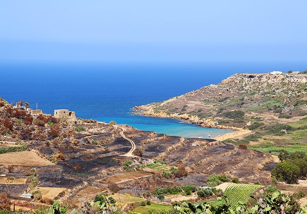 A view of the edge of the island of Gozo. Dry, tiered landscape tumbles into the blue Mediterranean Sea.