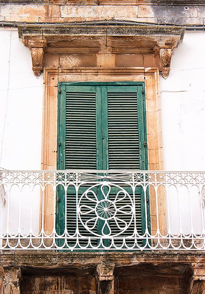 A close up of a green shutter on a white building with a decorative iron railing.