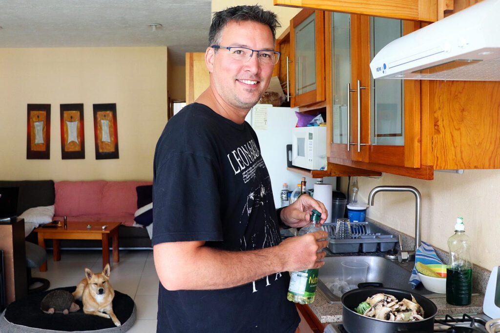 Gavin preparing dinner at our apartment in Ajijic. He is standing by the stove ready to add oil to a pot full of fresh vegetables. There is a sofa in the background and our dog on his bed. Cooking for ourselves is one way we spend less while traveling.
