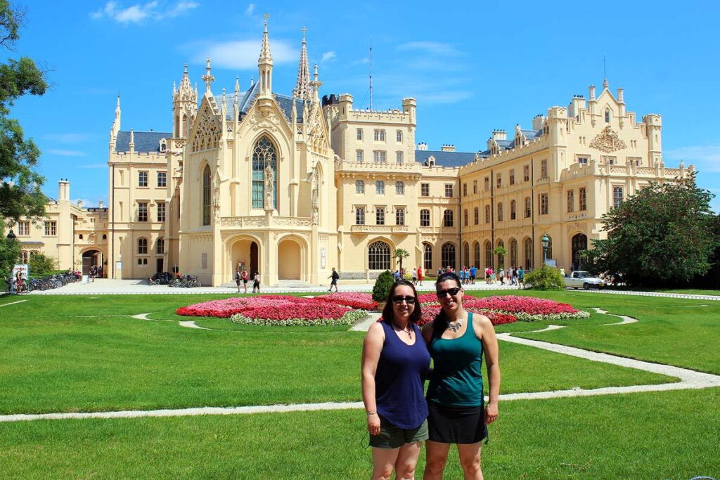 Beautiful Neo-Gothic castle in Lednice, Czech Republic. Robyn and her sister are standing in front of the green grass and colorful red and pink flowers. A great tip to travel more spend less is to travel with a companion to lower overall costs.