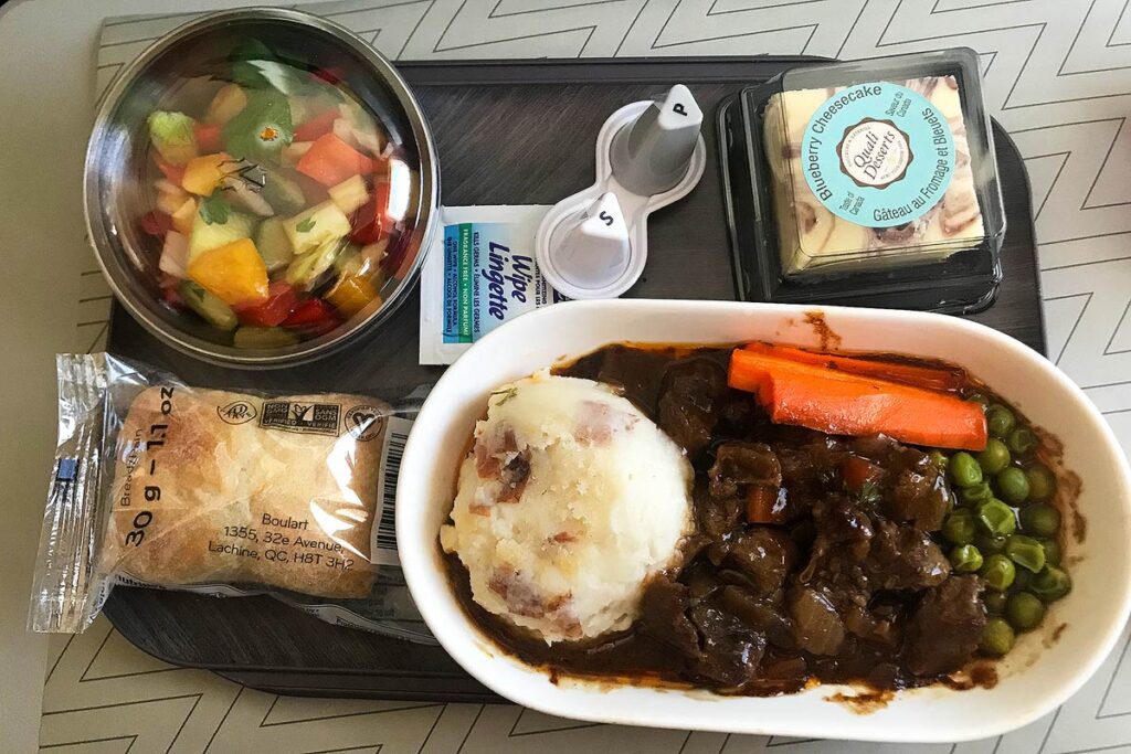 WestJet airplane dinner in premium economy. The main meal on the food tray is beef with mashed potatoes, carrots and peas in gravy. There is a desert in the top right corner opposite fruit salad, and a bun in a wrapper. If you want to travel for less, bring food on your flight.