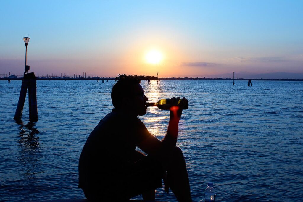 Gavin taking a sip from a bottle of wine on a small dock in Venice, Italy. The sun is setting creating a beautiful pink sky. The water is super blue and he is silhouetted. Buying wine at a local shop is cheaper than in a restaurant, and a great way to travel for less.