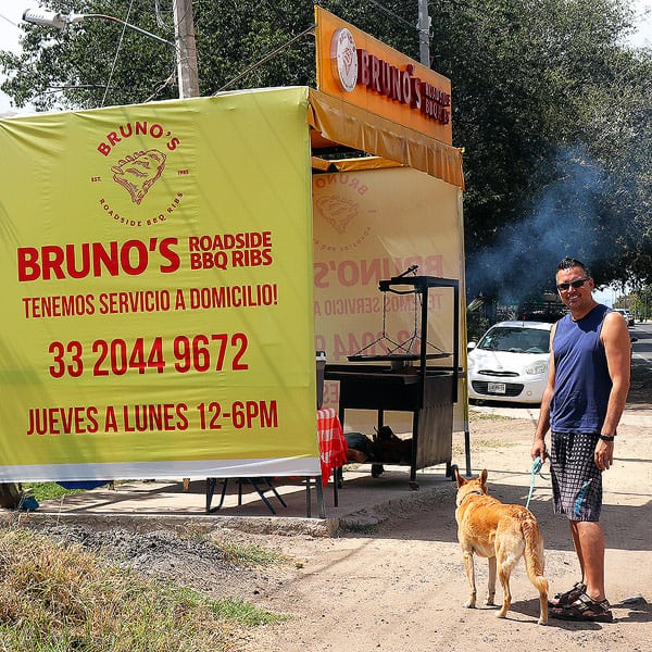 Gavin standing with our dog Leonardo in front of Bruno's Roadside BBQ Rib stand in Ajijic, Mexico. There is a large black BBQ and small table with a red and white tablecloth under a yellow tent. A great tip to travel more and spend less is to eat where the locals do.