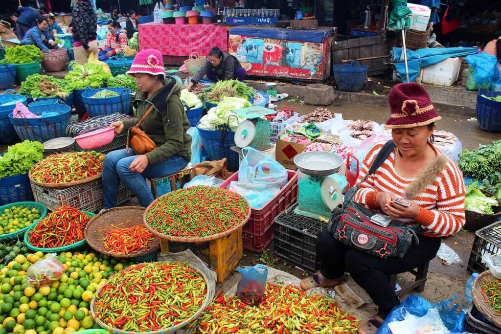 Morning market in Laos with women seated next to huge buckets of overflowing colorful peppers, limes, lettuce and other vegetables. A great tip to travel more spend less is to shop at local markets.