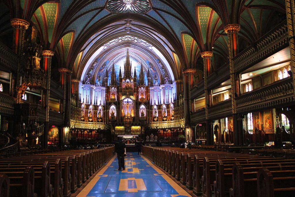 The stunning blue interior of the Basilica of Notre Dame in Montreal, Canada. The ceilings are a green blue and decorative wooden beams and benches fill the space. The alter is gothic style with blue background lighting. Most churches around the world are free to visit, and excellent way to spend less on travel.