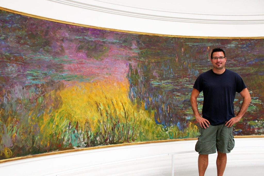 Gavin standing in front of a beautiful Monet painting in the Musee d'Orsay in Paris. The painting takes up the entire wall and showcases the waterlilies in purples, pinks and yellows. If you want to know how to travel for less, then visit a museum on the free day.