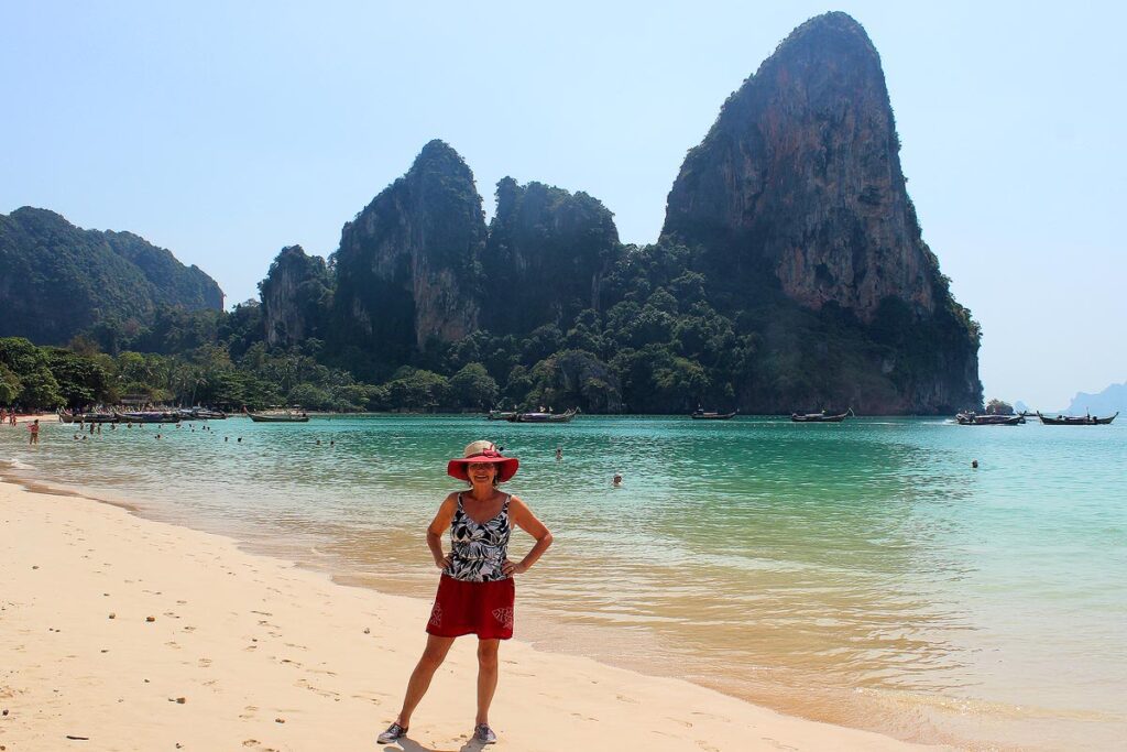 Gorgeous turquoise water and karst mountains of Railay Beach in Thailand. A lady wearing a black and white bathing suit with a red skirt & hat standing & smiling with hands on her hips. Creating a budget helps us spend less money while travelling.