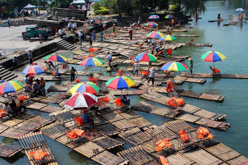 A chaotic scene of bamboo river boats in Yangshuo, China. There are rainbow umbrellas on half of the boats, along with simple chairs holding orange life vests. If you want to travel more spend less, then take local transportation.