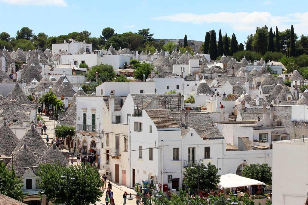 The white-washed town of Alberobello from a viewing point. Nearly every building is in the traditional trulli style with conical pointed roofs. There are green trees in the distance.