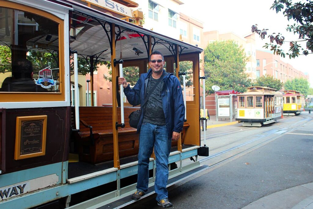 Gavin on a traditional cable car in San Francisco. There are 2 other cable cars in the distance. He is wearing jeans and a blue coats because it is spring. Taking public transportation is a great way to spend less on travel.