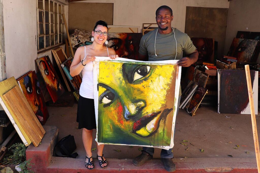 Robyn smiling and holding a beautiful large portrait painting with a local artist in Zimbabwe. They are in his workshop and you can see several other portraits leaning against the wall. Stick to one souvenir to help you spend less while traveling.