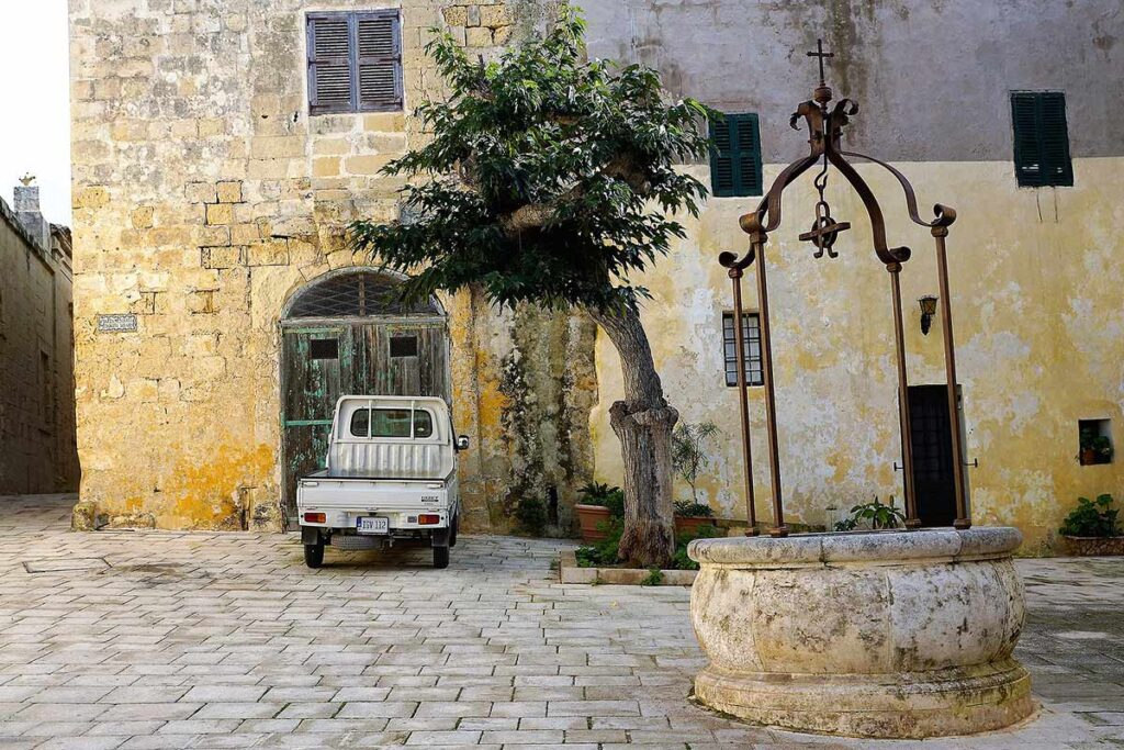 Quiet piazza with one tree, a well, and a single old truck in front of a doorway in Malta.
