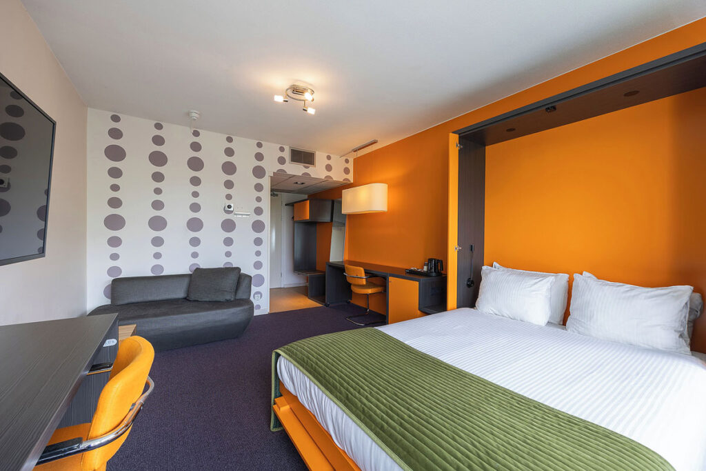 A modern room with orange walls, purple carpet and a large white bed with green cover. There is a couch, desk and chair in this suite from Amrath Airport Hotel, one of the best Rotterdam hotels with terrace near the airport.