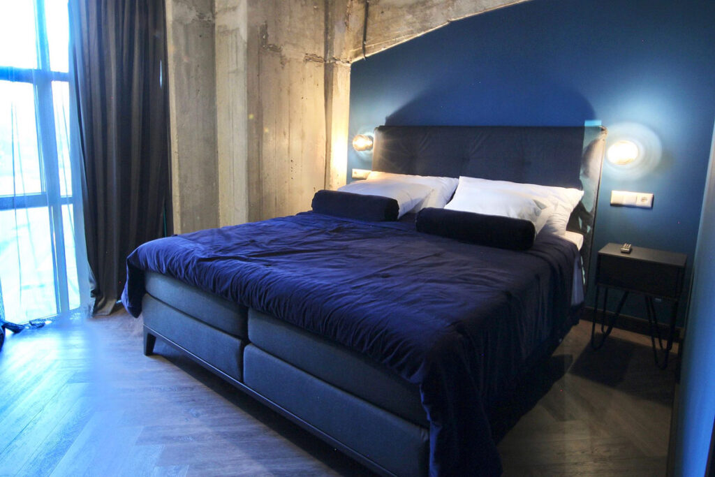 A room with concrete & blue colored walls and drapes at the Bed Bites & Business Rotterdam Hotel. There is a large bed with blue duvet, and large windows.
