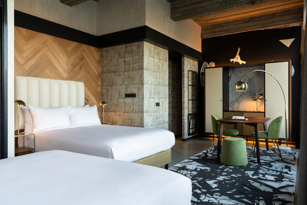 A spacious suite with two large beds at Fletcher Boutique Hotel Slaak-Rotterdam. There are exposed beams, textured walls, a desk, green sitting chair and black and white rug.