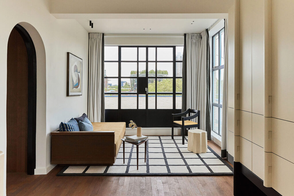 A contemporary living area in the Morgan & Mees Rotterdam hotel. The far wall is made of black paned windows and looks onto the city and river. There is a single bed, chair and black and white rug decorating the space.