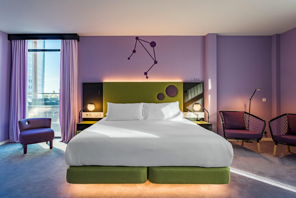 A suite with purple walls and a lime green headboard with purple circles for the comfortable queen bed at Room Mate Bruno, one of the best Rotterdam hotels with balcony. There is a purple chair in the right corner and a tall window with water views.