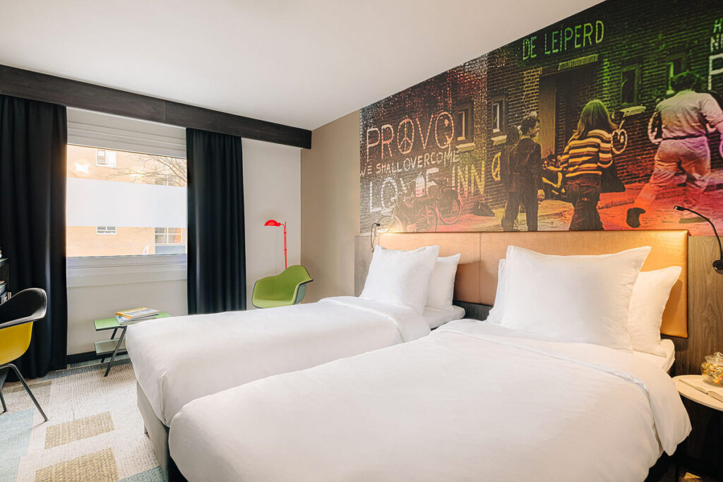 A room with two beds with white linens, a green chair and colorful, artsy photo as wallpaper. The Savoy Hotel is one of the best Rotterdam hotels with balcony in near nightlife.