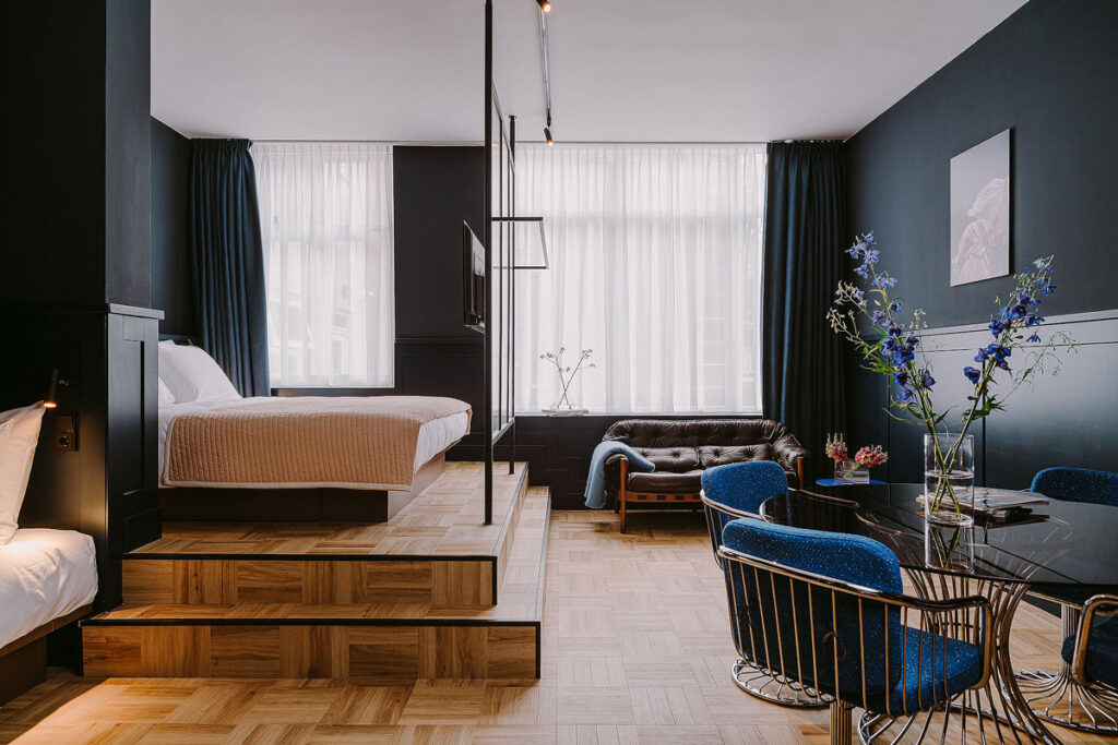 A beautiful suite with black walls, white curtains, a raised platform bed, a sofa, 2 blue chairs and wood flooring at the Supernova Hotel, one of the best Rotterdam hotels with balcony in the Centrum.