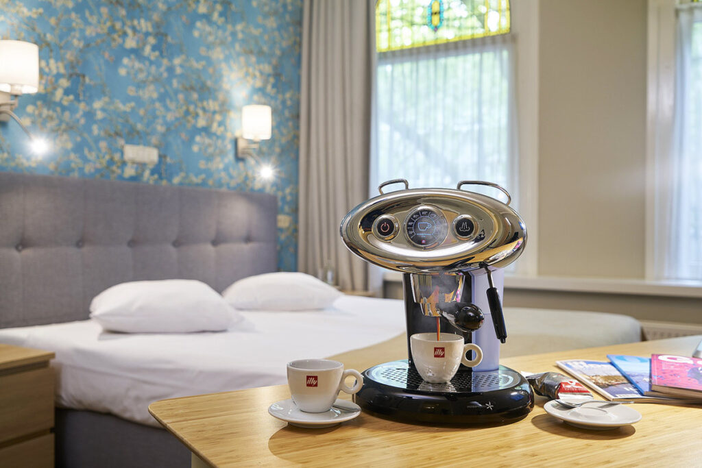 Close up of an Espresso machine in a suite with large windows, blue wallpaper and a large bed with grey headboard.