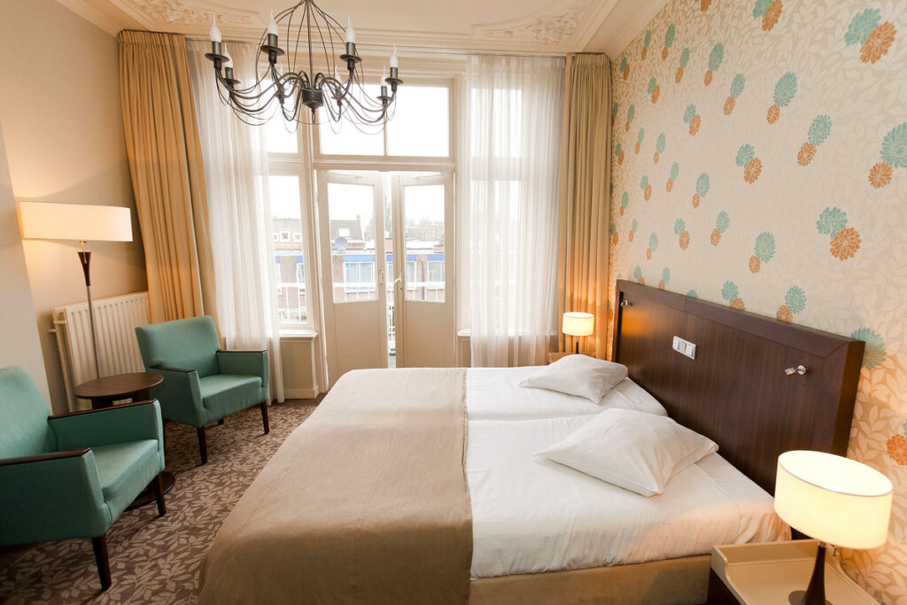A romantic suite at the Hotel Van Walsum, a Rotterdam hotel with balcony in the city centre. The room is bright and airy with open doors leading to the private terrace. There is soft colored decor, a chandelier above a large bed and two green chairs in the corner.