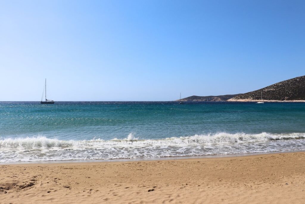 A picture of Platis Gialos, one of the best Sifnos beaches to visit. The water is aqua blue and vibrant turquoise and the sand is golden. There are mountains in the distance on the right and a single sailboat on the left.