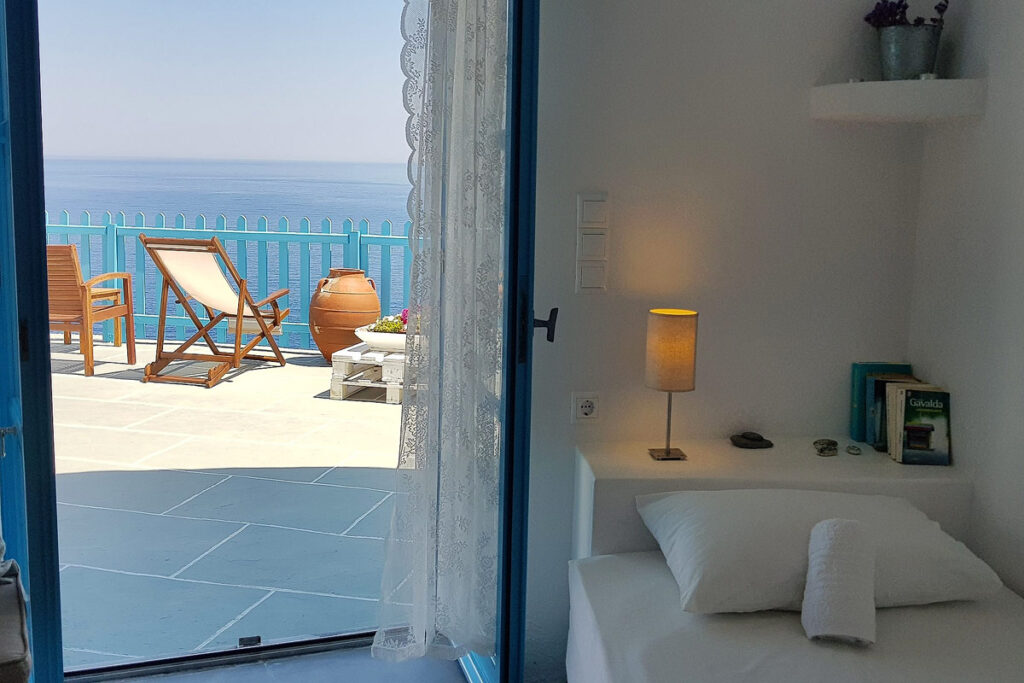 A single bed on the right with a small lamp on a ledge behind at the Aegean Eye Apartments in Seralia. The patio door is open to the furnished balcony with incredible sea views.
