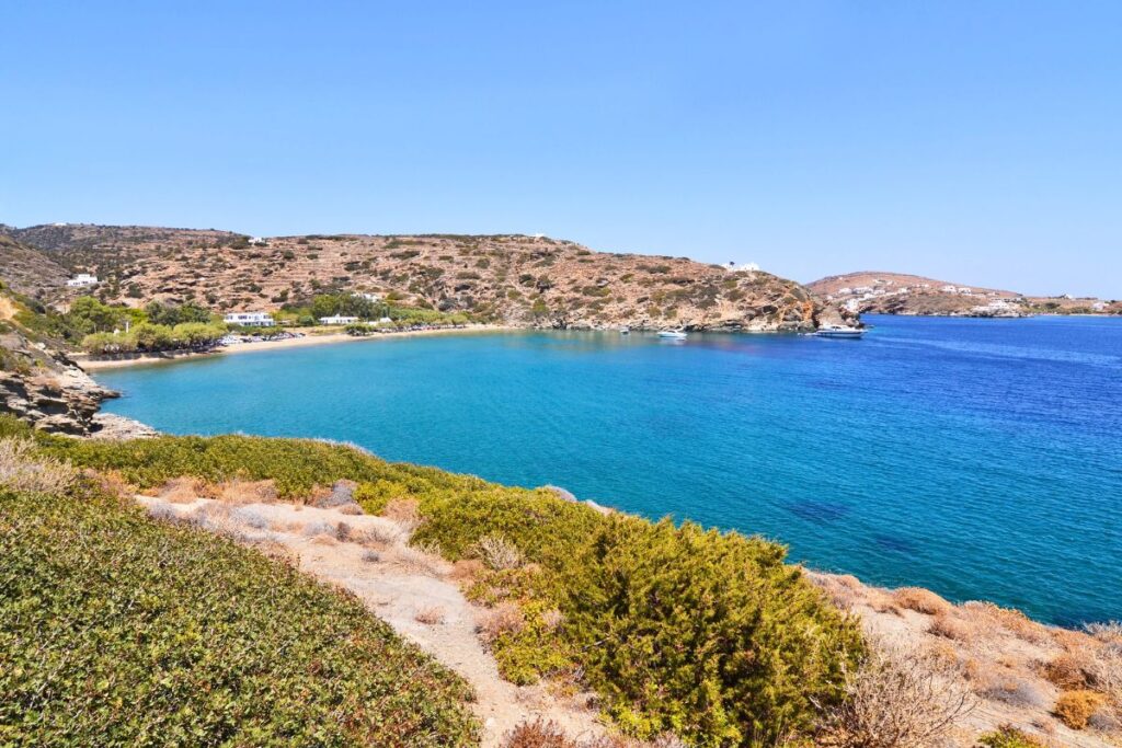 A panoramic view of Apokofto Beach with its bright, turquoise water and surrounding mountains. In the foreground is the top of a hill with green grass and shrubs, as well as a walking path.