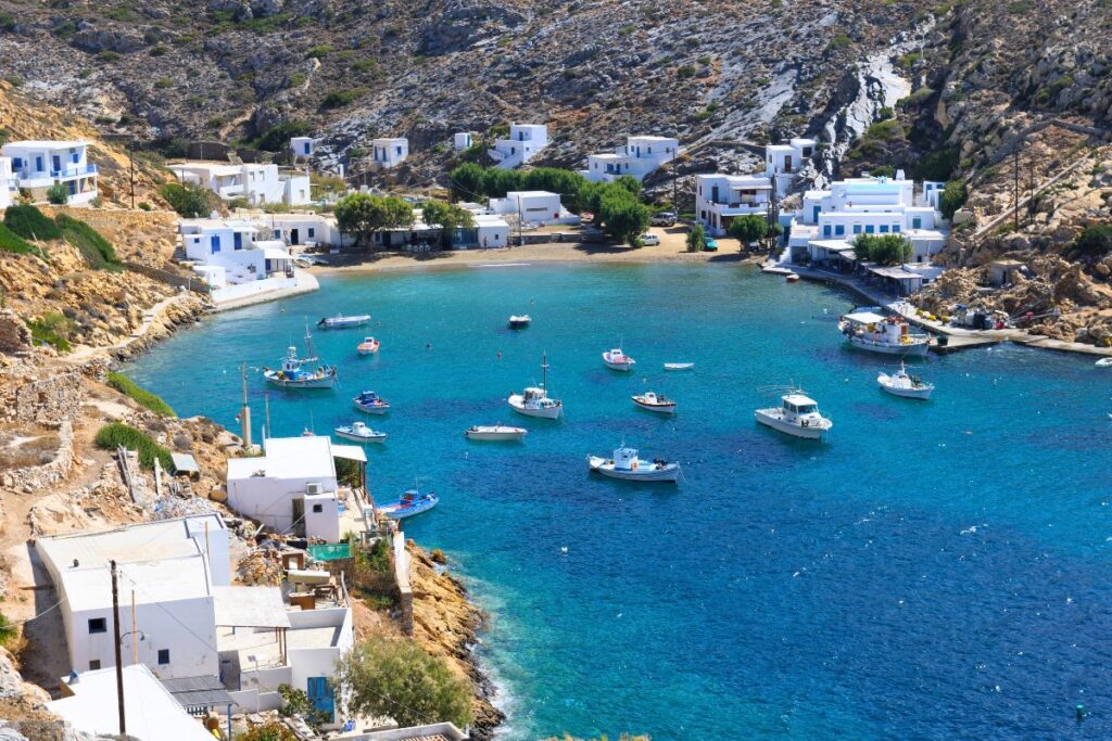 A bird's eye view of Cheronissos Beach, the most northern Sifnos beach. Several boats are anchored in the water, with many white buildings surrounding the bay. The water is turquoise and the beach has a few trees.