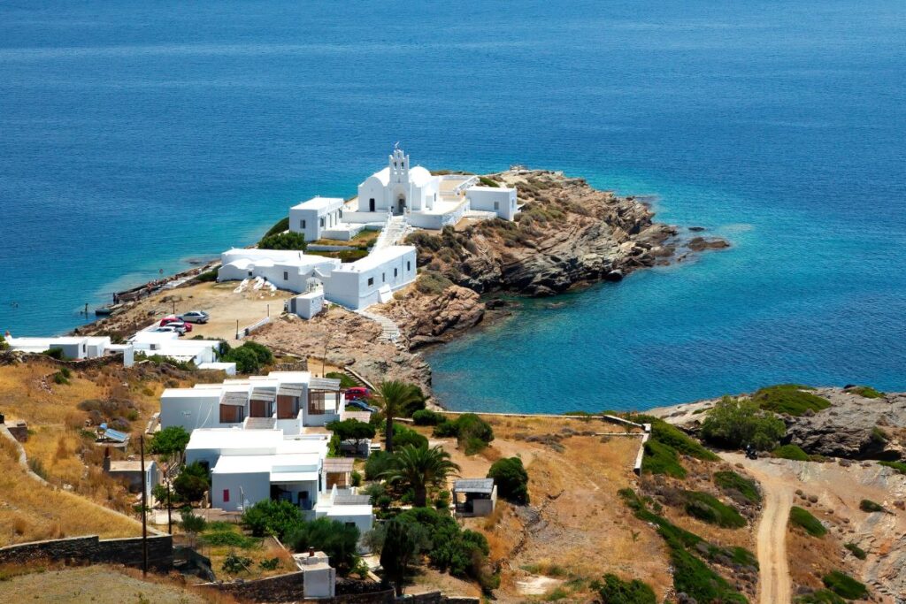 A bird's eye view of the iconic white Chrisopigi Monastery and surrounding area. There are a few white buildings sitting on the rocky landscape facing the deep aqua blue sea.