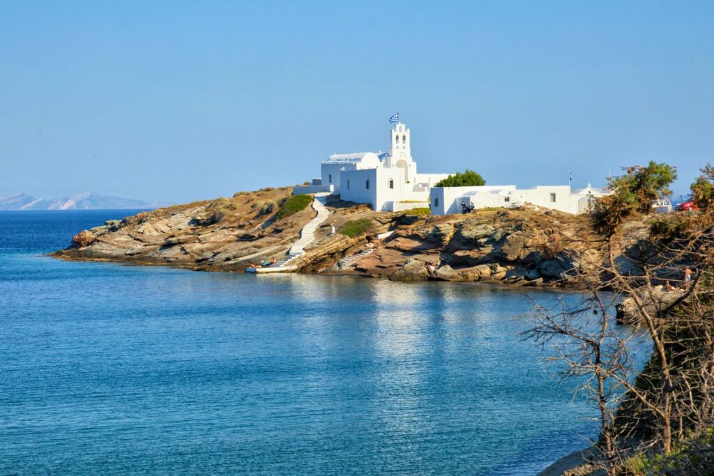 The iconic white Chrisopigi Monastery on a rocky jut out surrounded by beautiful water. Not a typical Sifnos beach, there are white stairs leading to the flat rocks below where people swim.