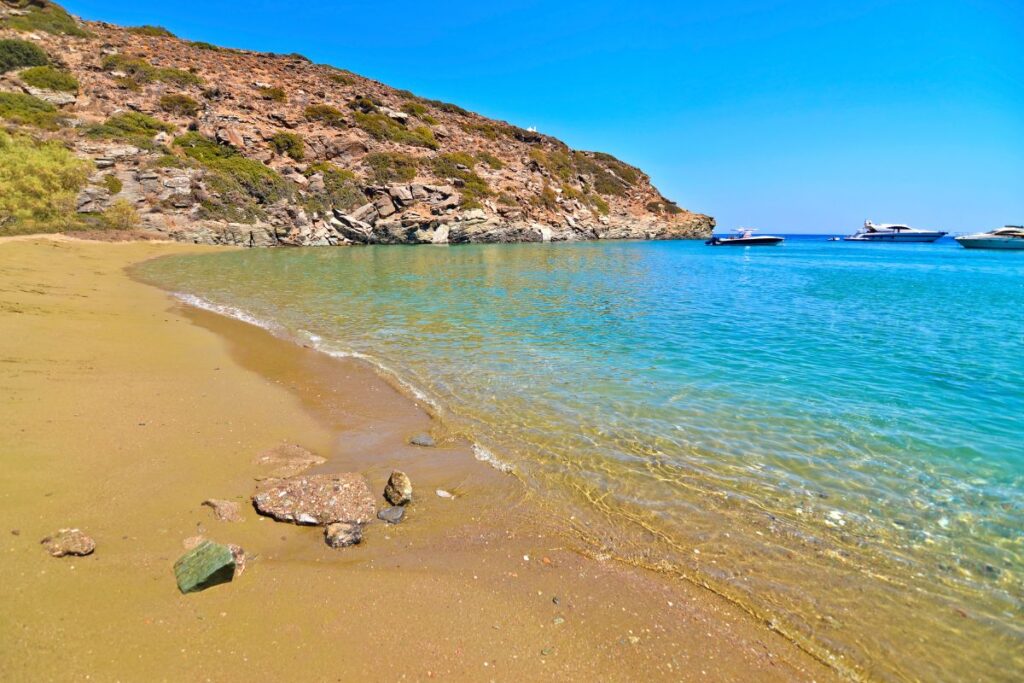 A beautiful view of Dialiskari, a less visited beach on Sifnos. There is crystal clear water, soft sand a mountain in the distance and a couple speedboats in the water. You can only access this beach by boat.