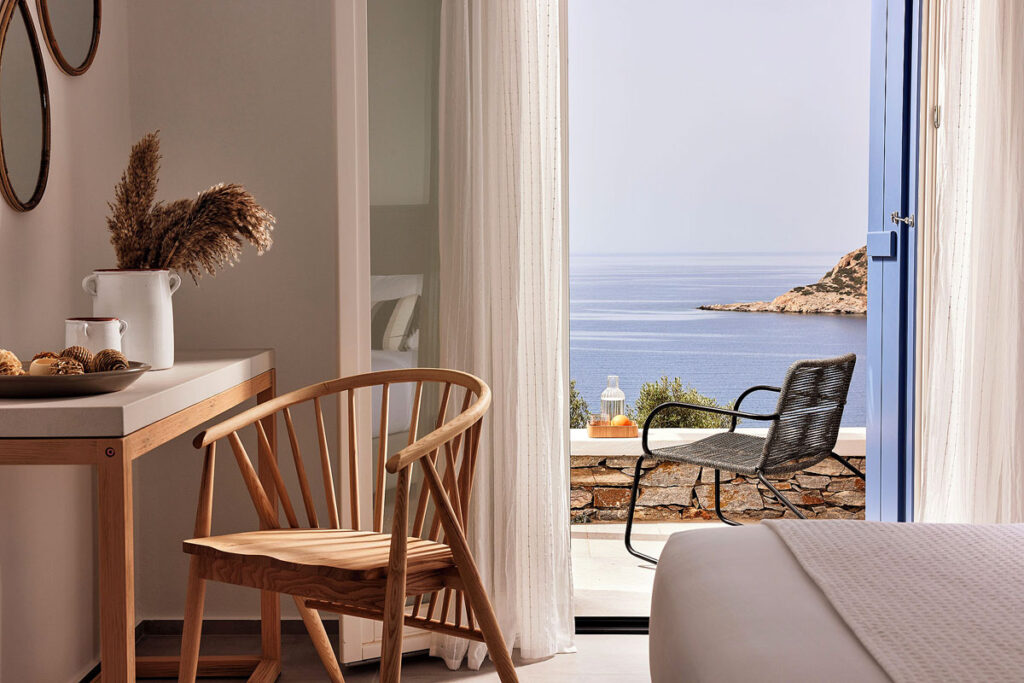 A gorgeous sea view from an open patio door at the Eutopia Suites Sifnos. There is a chair and bed in the foreground but the image draws your eyes out to the beautiful terrace and view.