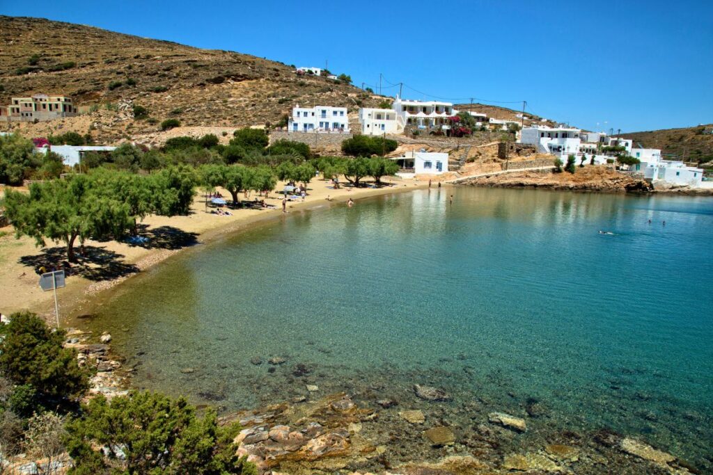 Lush, green trees sit on the shoreline of Faros beach, one of the best Sifnos beaches to visit. The water is clear and very calm, with some rocks in the foreground. There are a couple of white buildings in the distance sitting on the hill under the blue sky.