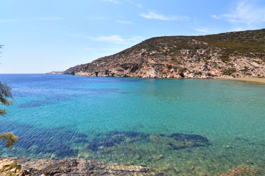 The calm, turquoise water of Fassolou, a beautiful beach in Sifnos. You can see rocks in the foreground under the clear water and a mountain in the distance with no houses.