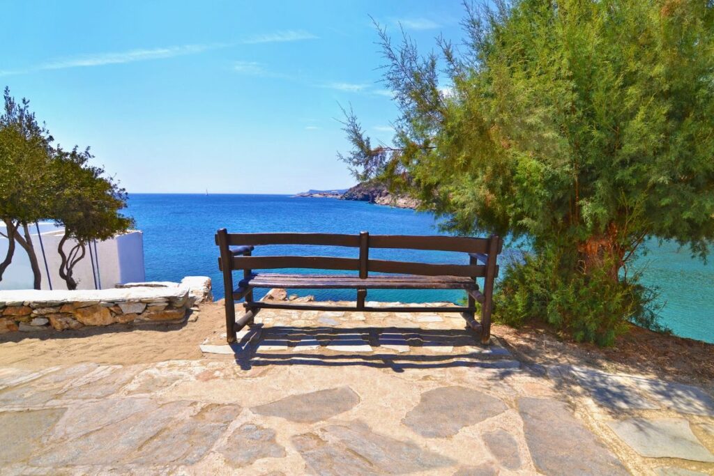 A single wooden bench beside a lush green tree looking out onto the deep blue Aegean Sea.