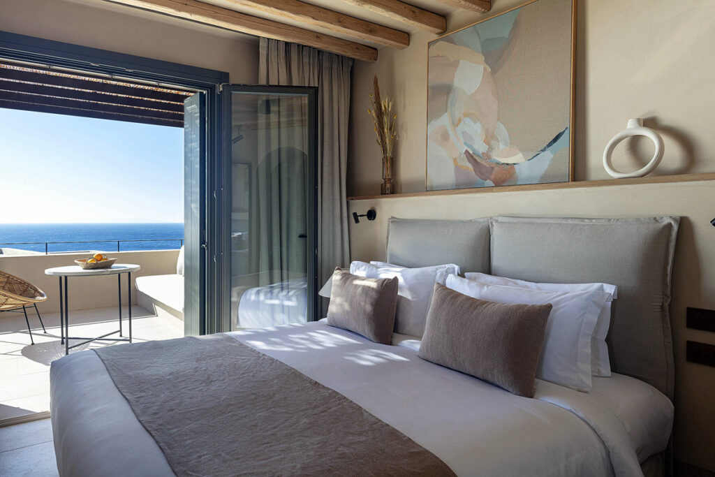 A beautiful suite at Nos Hotel & Villas. There is a large white bed with beige pillows, a painting on the wall and wooden beams. The patio doors are wide open leading to the furnished private terrace overlooking the sea.
