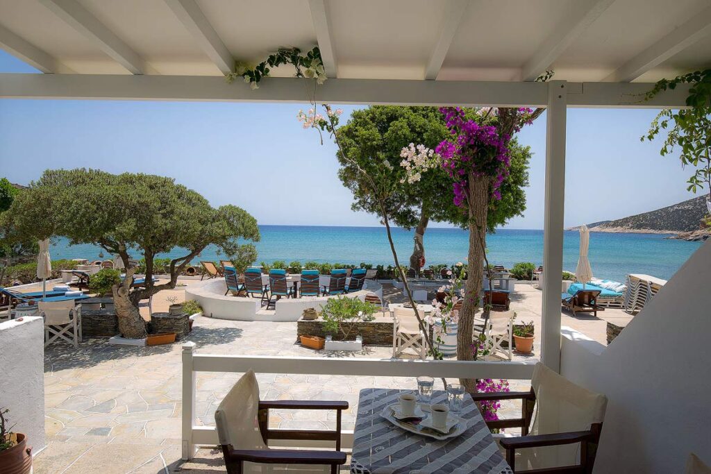 A patio with 2 chairs and table at the Pension Ageliki, one of the few Sifnos hotels on the beach. There are sun loungers, pink flowers and trees on the terrace with the beautiful turquoise water in the distance.