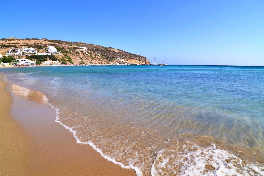 A close up ant's eye view of the beautiful clear turquoise water in Platis Gialos, one of the best Sifnos beaches to visit. You can see a rocky mountain in the distance with a few buildings. The sky is bright blue above.