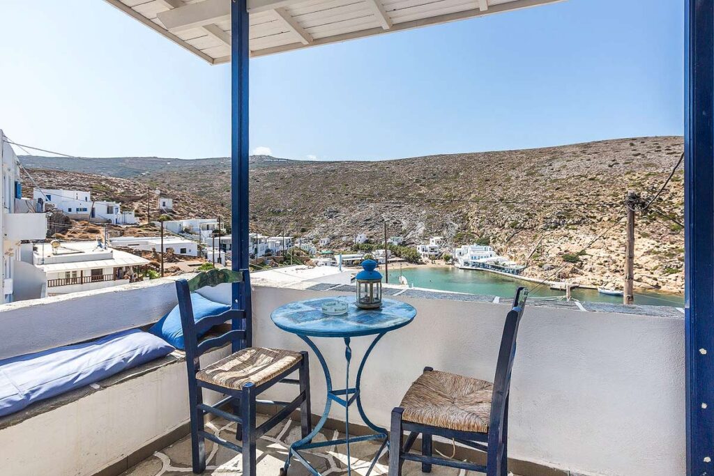 Two wicker chairs and a small round table on a furnished balcony overlooking Cheronissos, the most northern Sifnos beach to visit. You can see the small village, sandy beach and mountains in the background.