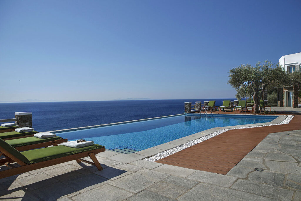 The gorgeous pool at Selana Suites overlooking the deep blue Aegean Sea below. There are 4 sun loungers with green cushions and white towels folded at one end and 4 others at the far end.